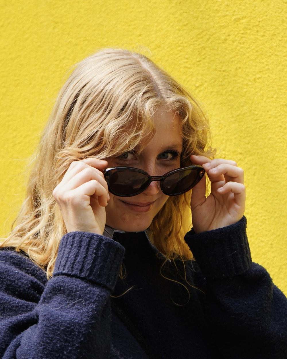 Portrait d'une femme blonde qui regarde l'objectif par-dessus ses lunettes de soleil, mur jaune